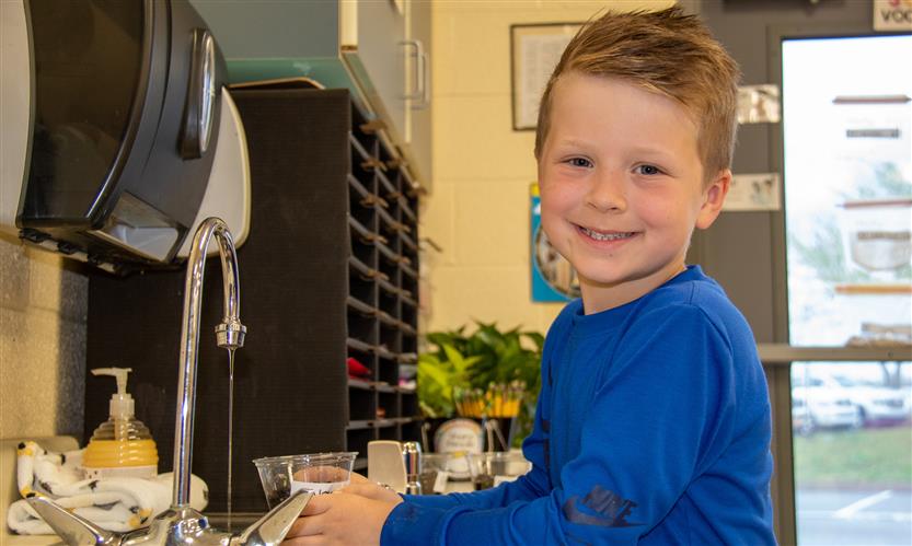 A boy wearing a blue shirt puts water into a plastic cup filled with dirt.